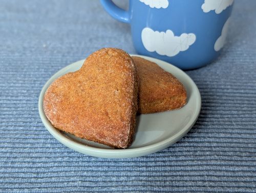 Zwei herzförmige Topfenteigkekse auf einem kleinen Teller auf einem blauen Tischuntersetzer mit einer blauen Tasse im Hintergrund